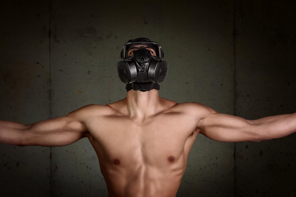 A shirtless man poses against a concrete wall wearing a gas mask